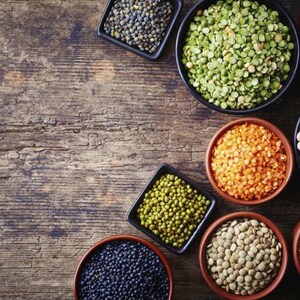 May include: A variety of dried legumes in small bowls on a wooden surface. The legumes include green lentils, black lentils, red lentils, chickpeas, split peas, and yellow split peas.