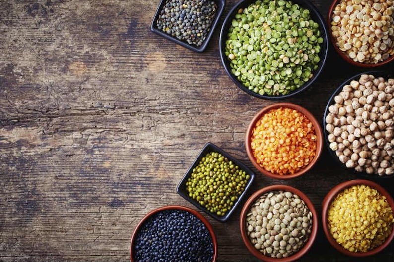 May include: A variety of dried legumes in small bowls on a wooden surface. The legumes include green lentils, black lentils, red lentils, chickpeas, split peas, and yellow split peas.