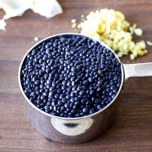May include: A close-up of a metal measuring cup filled with black lentils. The cup is sitting on a wooden surface.
