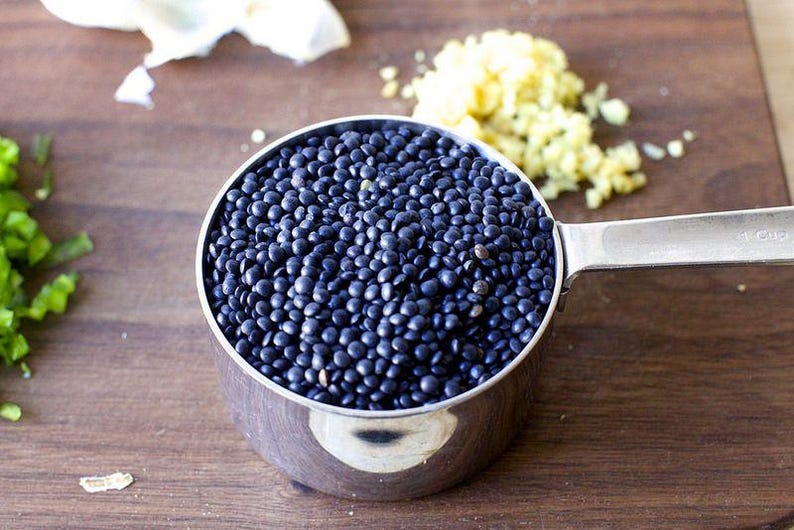 May include: A close-up of a metal measuring cup filled with black lentils. The cup is sitting on a wooden surface.