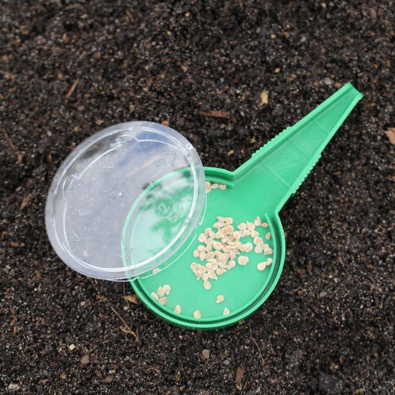 K&ouml;nnte beinhalten: Gr&uuml;ner Plastik-Saatgutspender mit einem durchsichtigen Deckel. Der Spender ist mit kleinen, hellbraunen Samen gef&uuml;llt.
