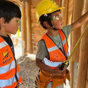 May include: Two children wearing construction vests, hard hats, and safety glasses. One child is using a measuring tape. The vests are orange with reflective stripes and logos. The scene takes place in a wooden structure, suggesting a construction or building theme.