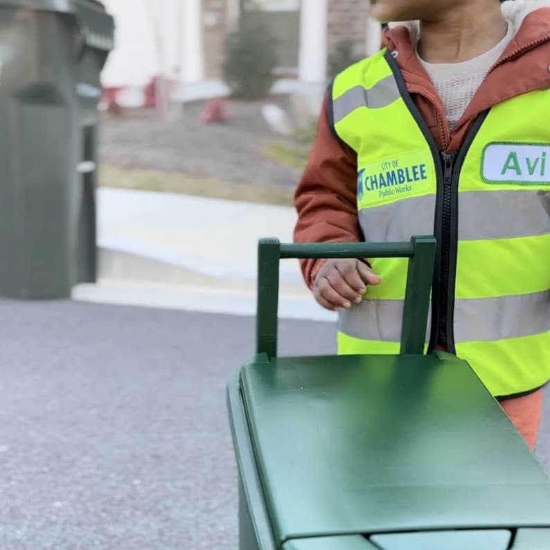 May include: A young person wearing a green and yellow safety vest with the text "City of Chamblee Public Works" and a name tag that says "Avi". The person is holding a green recycling bin with a handle.