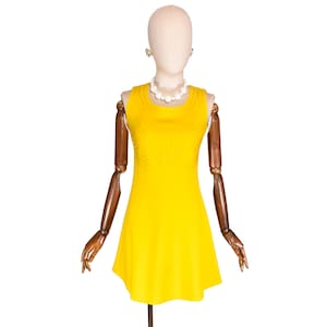 Puede incluir: Un vestido amarillo brillante sin mangas sobre un maniquí. El vestido presenta un diseño geométrico en el corpiño y una falda acampanada. El maniquí está adornado con un collar de perlas y pendientes.