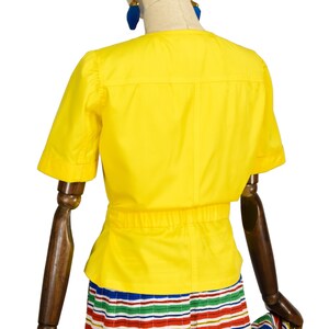 May include: A bright yellow short-sleeved blouse featuring a round neckline, a horizontal seam across the upper back, and a gathered elasticized waistline. The garment is displayed on a mannequin, paired with a multi-colored striped skirt.