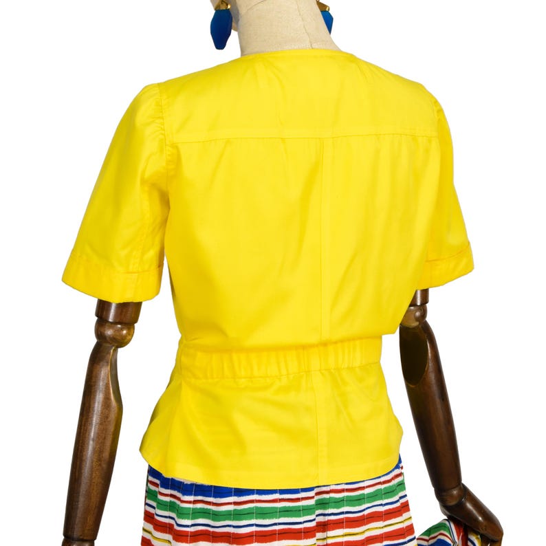 May include: A bright yellow short-sleeved blouse featuring a round neckline, a horizontal seam across the upper back, and a gathered elasticized waistline. The garment is displayed on a mannequin, paired with a multi-colored striped skirt.
