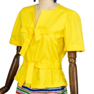 May include: A bright yellow short-sleeved blouse featuring a round neckline with a front slit, a tie detail at the chest, and a cinched waist with a matching tie belt. The garment has puffed sleeves and is displayed on a wooden mannequin.