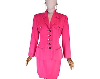 May include: A bright pink skirt suit displayed on a mannequin. The suit features a fitted blazer with silver buttons and two front pockets, paired with a matching knee-length skirt. The blazer has long sleeves and a classic collar. The mannequin is wearing black earrings.