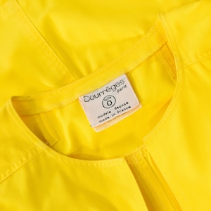 May include: A bright yellow garment featuring a round neckline and visible stitching along the collar. A white fabric label is sewn into the inner neck, displaying the text Courr&egrave;ges paris, taille 0, modele depose, and made in france.