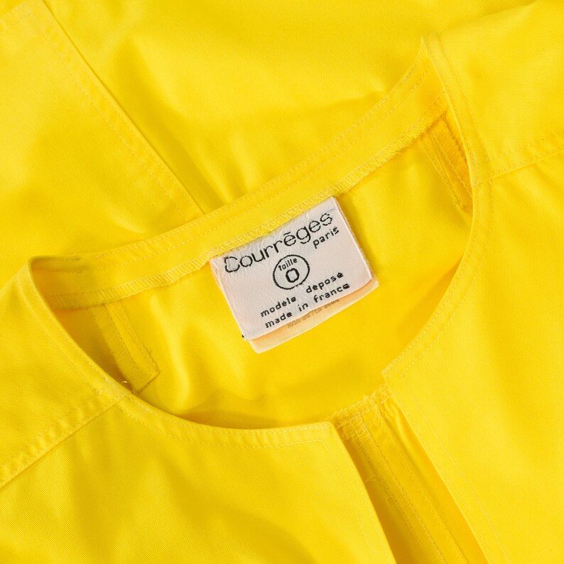 May include: A bright yellow garment featuring a round neckline and visible stitching along the collar. A white fabric label is sewn into the inner neck, displaying the text Courr&egrave;ges paris, taille 0, modele depose, and made in france.