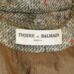 May include: Close-up of a tweed jacket collar with a label that reads "Ivoire de Balmain Paris." The jacket has a green and beige herringbone pattern with colorful flecks. The lining is a solid brown fabric.