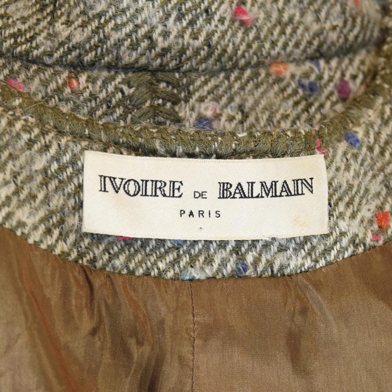 May include: Close-up of a tweed jacket collar with a label that reads "Ivoire de Balmain Paris." The jacket has a green and beige herringbone pattern with colorful flecks. The lining is a solid brown fabric.