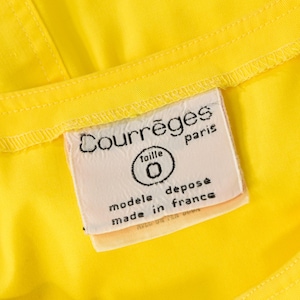 May include: A bright yellow garment featuring a white rectangular fabric label stitched onto the interior. The label displays the black text Courr&egrave;ges paris, taille 0, mod&egrave;le d&eacute;pos&eacute;, and made in france, with visible stitching along the edges.