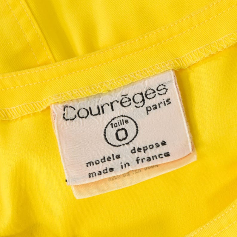 May include: A bright yellow garment featuring a white rectangular fabric label stitched onto the interior. The label displays the black text Courr&egrave;ges paris, taille 0, mod&egrave;le d&eacute;pos&eacute;, and made in france, with visible stitching along the edges.