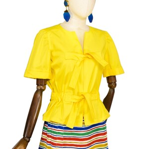 May include: A bright yellow short-sleeved blouse featuring a round neckline with a front tie closure and a cinched waist with a matching tie belt. The blouse is paired with a skirt featuring horizontal stripes in red, blue, green, and white.