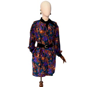 Op de afbeelding: Vintage jurk met bloemenprint met lange mouwen, een zwarte fluwelen kraag en bijpassende riem. De jurk heeft een levendig patroon van oranje, paarse en blauwe bloemen. De jurk wordt op een mannequin gepresenteerd.