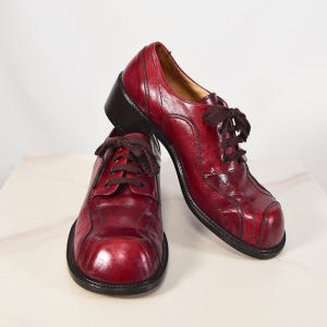May include: A pair of burgundy leather shoes with dark brown laces. The shoes have a rounded toe and a small heel. The stitching is visible, and the shoes appear to be vintage or retro style.