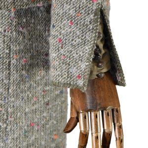 May include: Close-up of a tweed jacket sleeve. The jacket is a light green color with flecks of red, blue, and orange. The sleeve has a buttoned cuff and a contrasting olive green lining. The jacket is displayed on a wooden mannequin hand.