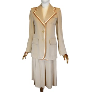May include: A beige skirt suit with a white and brown trim. The jacket has a notched collar and two button closure. The skirt is pleated.