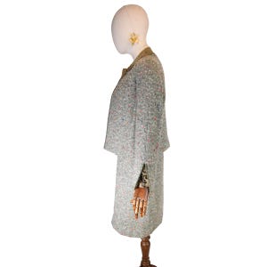 May include: A vintage tweed suit in a light gray with flecks of red, blue, and yellow. The suit includes a long jacket with a brown collar and a matching skirt. The outfit is displayed on a mannequin with a wooden arm and a neutral-toned head.