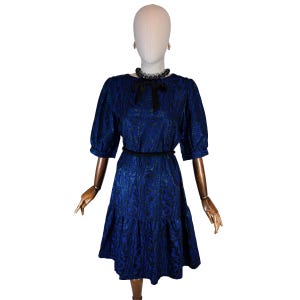 Op de afbeelding: Een blauwe jurk met een zwart patroon en korte mouwen wordt getoond op een mannequin. De jurk heeft een zwarte riem, een zwarte strik bij de halslijn en een rok met ruches. De mannequin draagt ook een zwarte kralenketting en oorbellen.