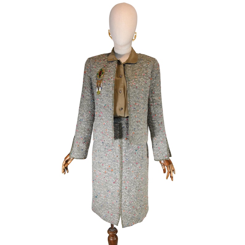 May include: A vintage tweed suit displayed on a mannequin. The suit includes a long coat and a skirt, both in a speckled gray fabric with flecks of pink, blue, and orange. A gold-toned brooch and earrings add a touch of elegance. The suit is paired with a tan blouse.