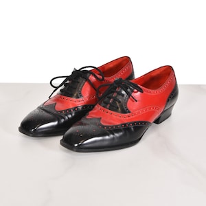 Puede incluir: Un par de zapatos Oxford bicolores de cuero rojo y negro. Los zapatos presentan un diseño clásico de ala con cordones negros y un tacón bajo. El cuero rojo está en la parte superior, mientras que la puntera y los detalles son negros.