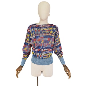 Puede incluir: Un suéter de punto con estampado geométrico azul, rojo, amarillo y blanco, con mangas largas y cuello de barco.