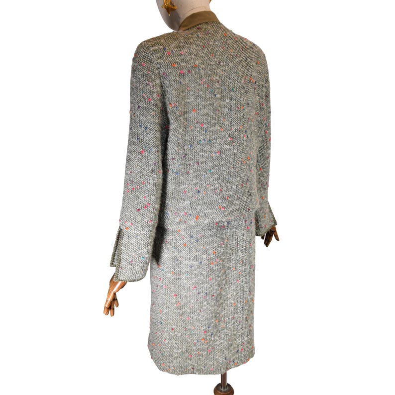 May include: A vintage tweed suit in a light gray hue, speckled with colorful flecks. The suit includes a jacket with a brown collar and a matching knee-length skirt. The jacket has long sleeves and a classic tailored fit.