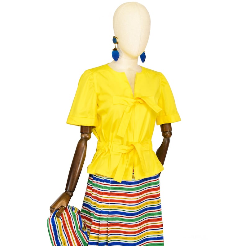 May include: A bright yellow short-sleeved blouse featuring a split neckline with a tie closure and a matching tie at the waist. The blouse is paired with a pleated skirt showcasing horizontal stripes in red, green, blue, and yellow, plus blue earrings.