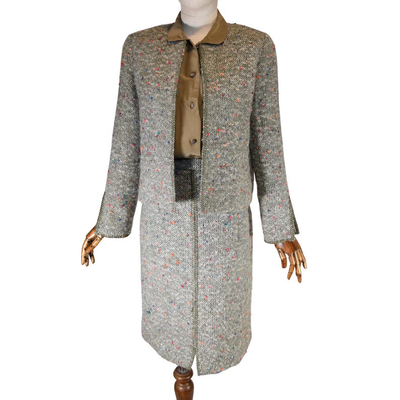 May include: A gray tweed skirt suit with a matching jacket and skirt. The jacket has a notched collar and long sleeves. The skirt is knee-length. The suit has flecks of color throughout. A tan blouse is visible underneath the jacket.