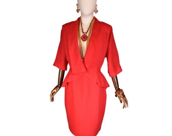 May include: A vibrant red two-piece suit displayed on a mannequin. The suit includes a blazer with three-quarter sleeves and a skirt. Gold-toned jewelry, including earrings, necklaces, and a bracelet, complements the outfit. The mannequin is on a wooden stand.