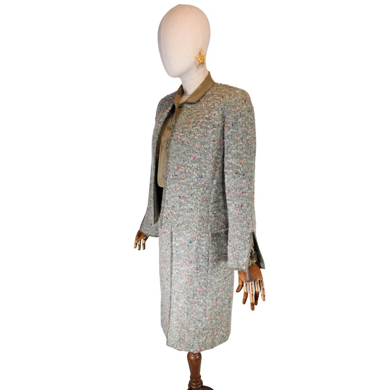 May include: A vintage tweed skirt suit in a light gray color with flecks of pink, blue, and orange. The suit includes a long jacket with a notched collar and a matching knee-length skirt. The outfit is displayed on a mannequin.