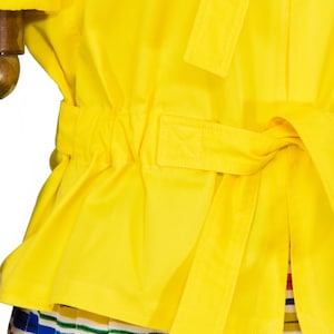 May include: A bright yellow jacket featuring a cinched waist with a matching fabric tie belt. The garment has a smooth texture and is worn over a skirt with horizontal stripes in blue, green, red, and white colors, visible at the bottom edge.