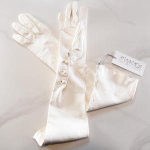 May include: A pair of long, white leather gloves with button details. The gloves are laid out on a light-colored surface, with a tag attached. The tag reads "ATTAVINTAGE".