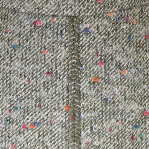 May include: Close-up of a textured tweed fabric in shades of gray and white, with a herringbone weave. The fabric is speckled with colorful flecks of pink, orange, blue, and teal. A vertical seam of olive green fabric runs down the center.