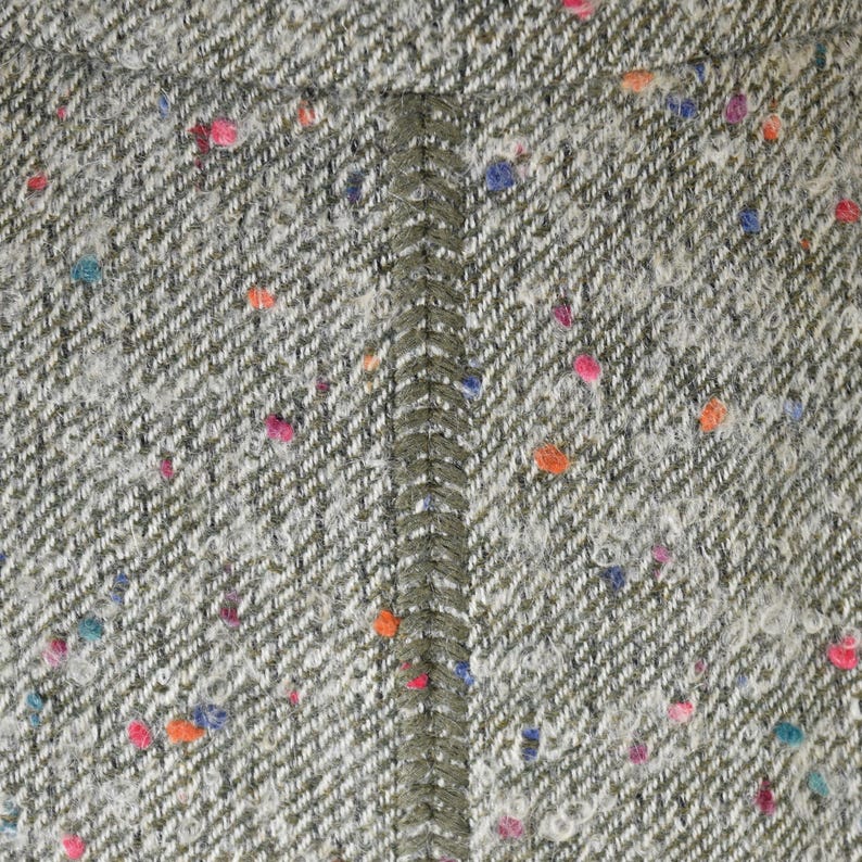 May include: Close-up of a textured tweed fabric in shades of gray and white, with a herringbone weave. The fabric is speckled with colorful flecks of pink, orange, blue, and teal. A vertical seam of olive green fabric runs down the center.