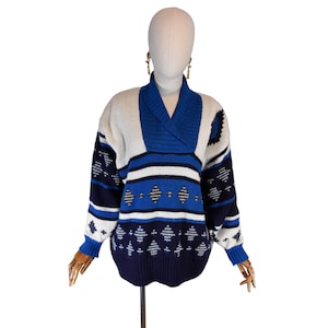 Könnte beinhalten: Ein Vintage-Pullover in Weiß, Marineblau und Blau, auf einer Schaufensterpuppe präsentiert. Der Pullover hat einen Schalkragen und ein geometrisches Muster mit Rauten und Streifen. Die Ärmel und der Saum sind gerippt.