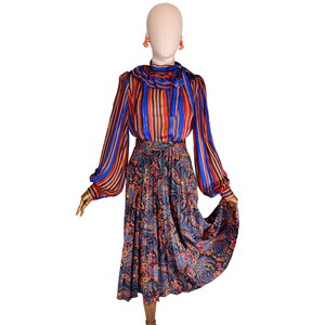 Può includere: Un completo vintage a due pezzi su un manichino. La parte superiore è una camicetta a righe verticali blu, arancioni e marroni. La gonna è lunga midi con un motivo paisley blu, arancione e nero. L'outfit ha maniche lunghe.