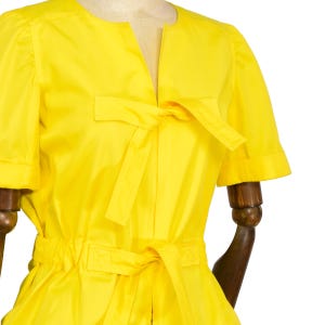 May include: A bright yellow short-sleeved blouse featuring a split neckline with a tie closure at the chest and a matching tie belt at the waist. The garment has a smooth finish and short sleeves with folded cuffs, presented on a mannequin.