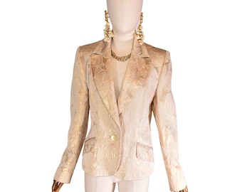 Pode incluir: Um blazer dourado com um padrão floral, exibido em um manequim. O blazer tem um fecho de um botão, lapelas entalhadas e dois bolsos frontais. O manequim também é adornado com joias douradas, incluindo um colar e brincos grandes.