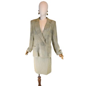 Può includere: Un tailleur gonna verde chiaro esposto su un manichino. La giacca doppiopetto e la gonna al ginocchio sono realizzate in un tessuto strutturato con un sottile motivo geometrico. L'abito presenta un risvolto a lancia e due tasche anteriori. Il manichino indossa orecchini dorati.