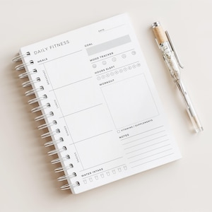 May include: A white Daily Fitness journal with a spiral binding, featuring sections for meals, mood tracking, and workouts. A clear pen with a marbled design and silver details lies next to the journal. Includes sections for water intake and notes.