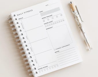 May include: A white Daily Fitness journal with a spiral binding, featuring sections for meals, mood tracking, and workouts. A clear pen with a marbled design and silver details lies next to the journal. Includes sections for water intake and notes.
