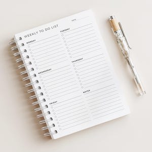 May include: A white spiral-bound weekly to-do list notepad with a silver pen. The notepad has a simple layout with sections for each day of the week and a notes section. The pen has a clear barrel with a decorative design.
