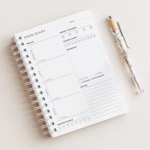 May include: A white spiral-bound food diary with the words "FOOD DIARY" at the top. The diary includes sections for meals, calories, mood tracking, and water intake. A clear pen with a silver clip and a cork-like top rests beside the diary.