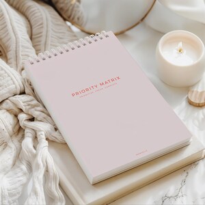 May include: A light pink spiral-bound notebook with the words "PRIORITY MATRIX" printed in red. The notebook is open on a white surface next to a lit candle in a white ceramic holder. A beige knitted blanket is draped nearby.