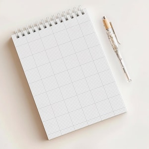 May include: A white spiral-bound notebook with a grid pattern and a silver pen with a marble-like design. The notebook is open, displaying the gridded pages. The pen is lying next to the notebook on a white surface.