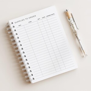 May include: A white spiral-bound notebook with the text "SUPPLIES TO ORDER" at the top. The notebook has columns for SKU, Item, QTY, Cost, and Order Date. A clear pen with a marbled design and a cork top rests beside the notebook.