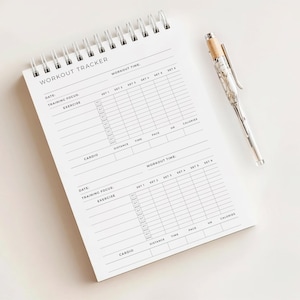 May include: A white spiral-bound workout tracker notebook with the words "WORKOUT TRACKER" printed at the top. A silver pen with a marble-effect design rests beside the notebook. The notebook has sections for date, focus, exercise, and workout time.